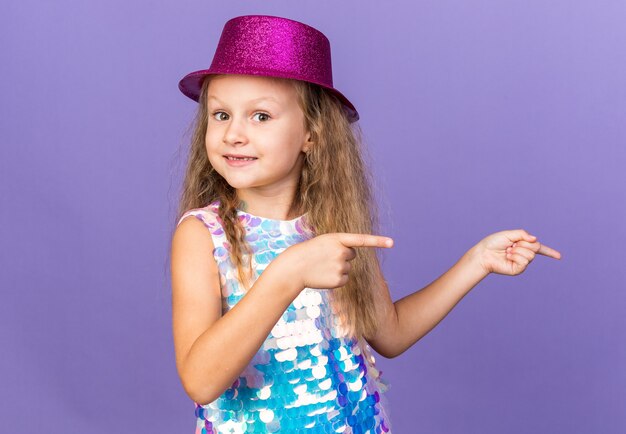 impressed little blonde girl with violet party hat pointing at side isolated on purple wall with copy space