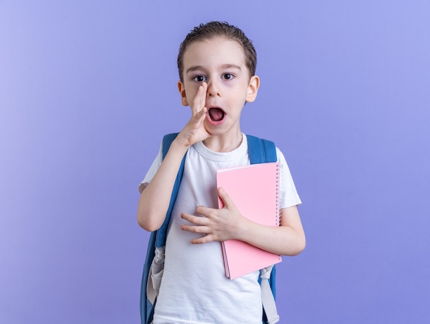 Impressed little boy wearing backpack holding note pad keeping hand near mouth looking at camera whispering isolated on purple wall with copy space