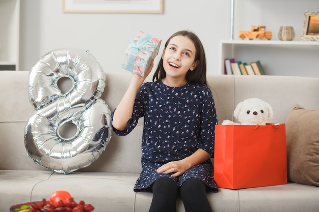 Free photo impressed little girl on happy woman's day holding present sitting on sofa in living room