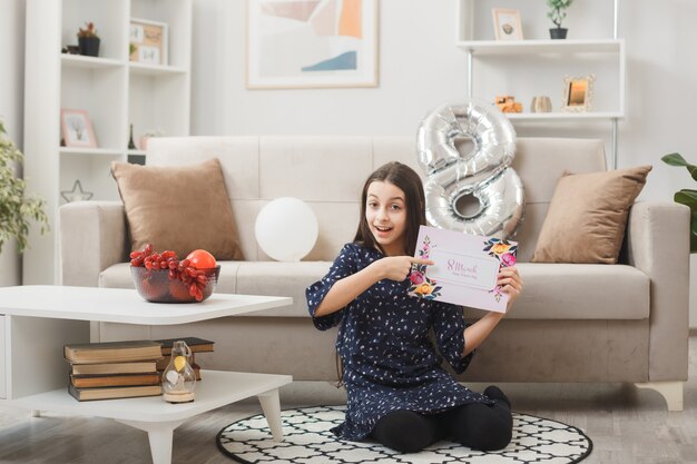 Impressed little girl on happy woman's day sitting on floor holding and points at postcard in living room