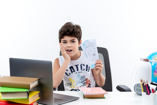 Impressed little schoolboy sitting at desk with school tools holding tickets and putting hand on cheek isolated on white background