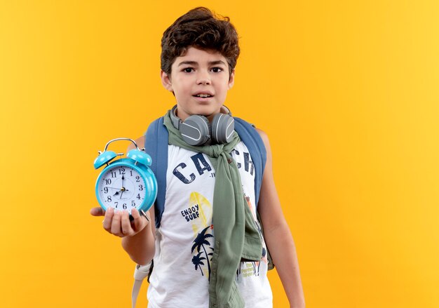 黄色の背景で隔離の目覚まし時計を保持しているバックバッグとヘッドフォンを身に着けている感動の小さな男子生徒