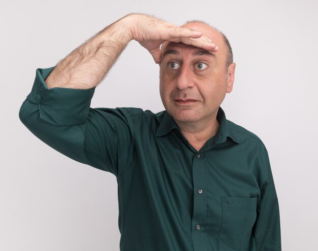Impressed looking at side middle-aged man wearing green t-shirt looking at distance with hand isolated on white wall