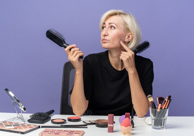 Impressed looking side young beautiful girl sits at table with makeup tools holding combs isolated on blue wall