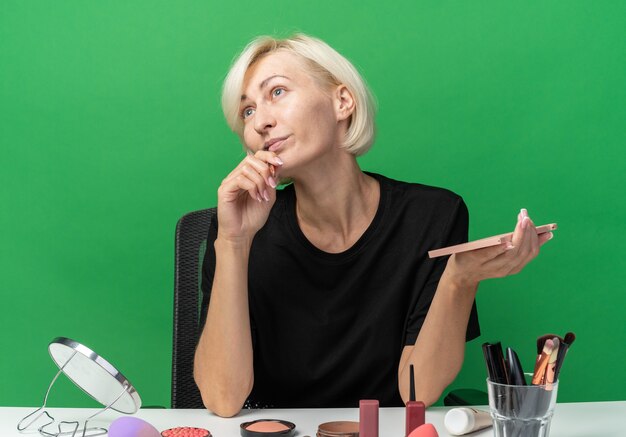 Impressed looking side young beautiful girl sits at table with makeup tools holding eyeshadow palette with makeup brush isolated on green wall