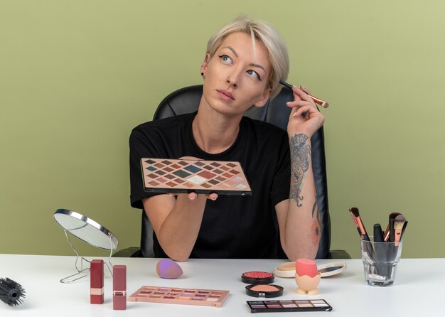 Impressed looking side young beautiful girl sits at table with makeup tools holding eyeshadow palette with makeup brush isolated on olive green wall