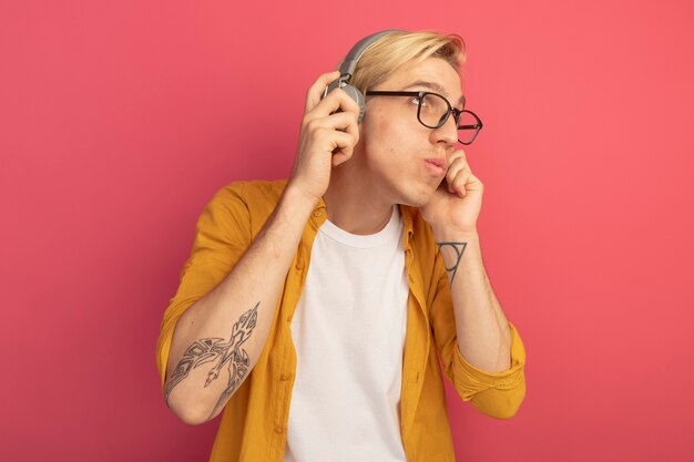 Impressed looking at side young blonde guy wearing yellow t-shirt and glasses with headphones isolated on pink