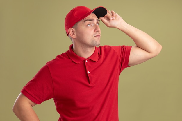 Impressed looking at side young delivery man wearing uniform and cap and holding cap isolated on olive green wall