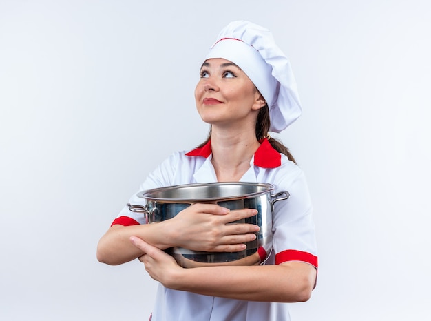 白い壁に隔離された鍋を保持しているシェフの制服を着ている印象的な側面の若い女性料理人