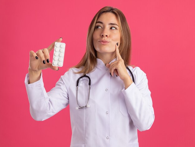 Impressed looking at side young female doctor wearing medical robe with stethoscope holding pills putting finger on cheek isolated on pink wall