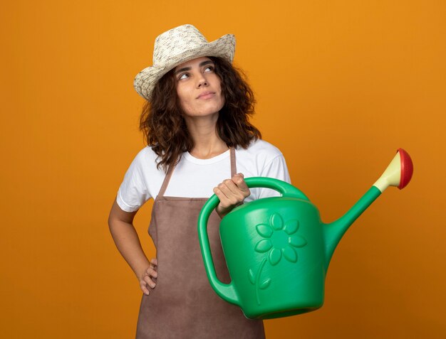 Impressed looking at side young female gardener in uniform wearing gardening hat holding watering can and putting hand on hip