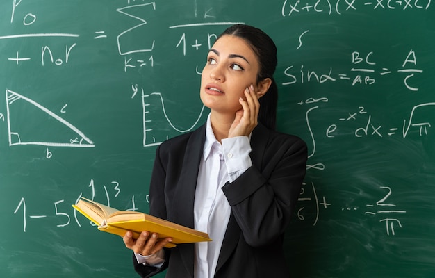 Impressed looking side young female teacher standing in front blackboard holding book putting hand on cheek in classroom