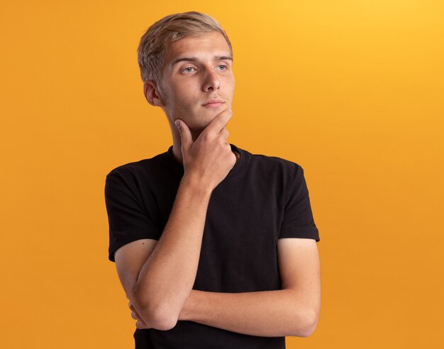 Impressed looking at side young handsome guy wearing black shirt grabbed chin isolated on yellow wall