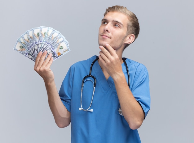 Impressed looking at side young male doctor wearing doctor uniform with stethoscope holding money putting hand under chin isolated on white wall