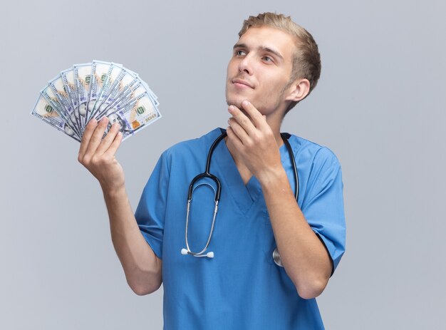 Impressed looking at side young male doctor wearing doctor uniform with stethoscope holding money putting hand under chin isolated on white wall