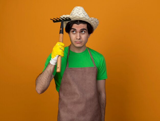 Impressed looking at side young male gardener in uniform wearing gardening hat with gloves holding rake