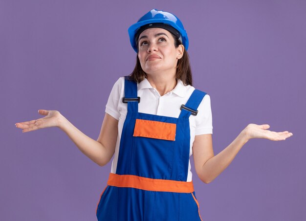 紫色の壁に分離された均一な広がりの手で若いビルダーの女性を見て感動