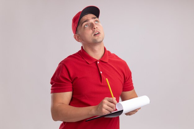 Impressed looking up young delivery man wearing uniform with cap holding clipboard with pencil isolated on white wall