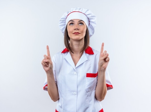 Foto gratuita impressionato guardando la giovane cuoca che indossa l'uniforme dello chef punta verso l'alto isolato sul muro bianco
