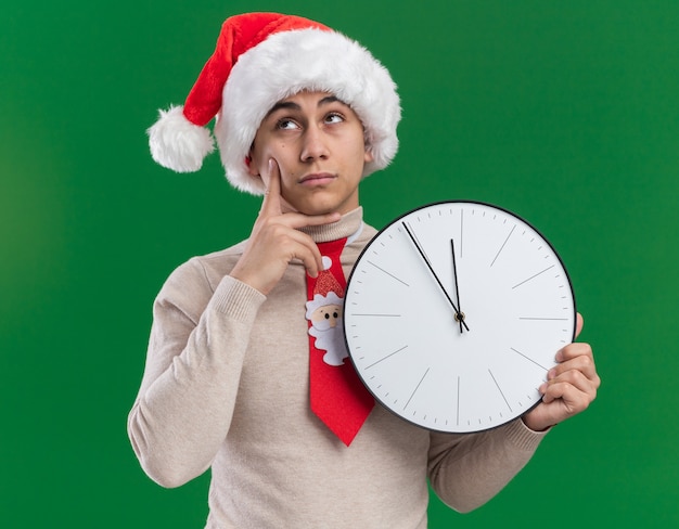 Под впечатлением от взгляда молодой парень в новогодней шапке с галстуком держит настенные часы и кладет палец на щеку, изолированную на зеленой стене