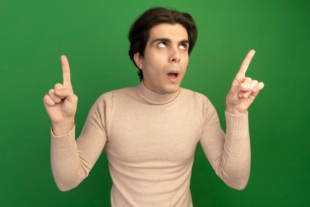 Impressed looking up young handsome guy points at up isolated on green wall