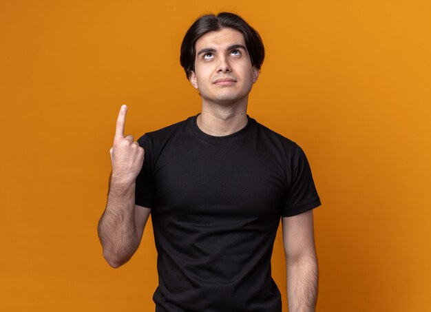 Impressed looking up young handsome guy wearing black t-shirt points at up isolated on orange wall