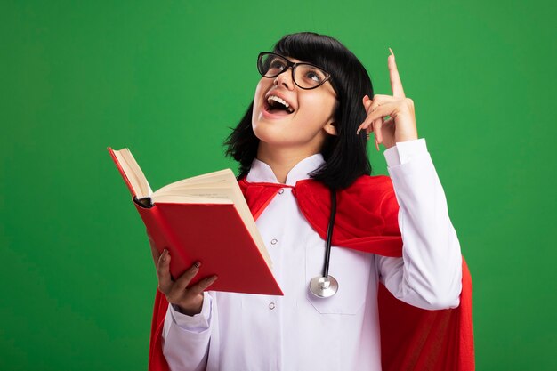 Impressed looking up young superhero girl wearing stethoscope with medical robe and cloak with glasses holding book points at up isolated on green wall