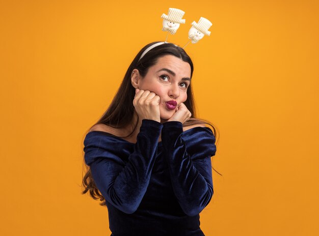 Impressed looking upyoung beautiful girl wearing blue dress and christmas hair hoop putting hands on cheeks isolated on orange background