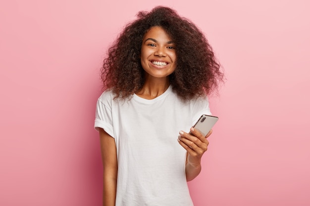 Impressed lovely Afro woman with luxurious curly hair, holds modern mobile phone, waits for call