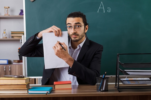 教室で学校の道具とテーブルに座っているペンで紙を保持している眼鏡をかけている感銘を受けた男性教師