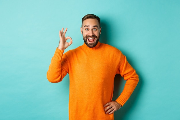 Impressed man recommending promo, showing okay sign and smiling amazed, approve and agree, standing over light turquoise wall.