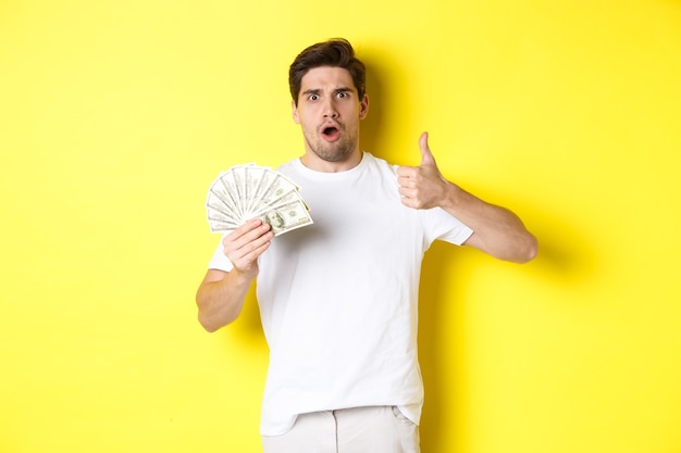 Impressed man showing thumb up, holding money credit, standing over yellow background.