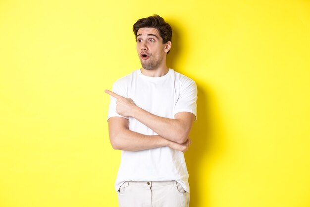 白いTシャツを着た印象的な男、プロモーションで左の指を見て人差し指、広告をチェック、黄色の背景に立って