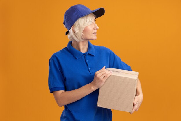 Impressed middle-aged blonde delivery woman in blue uniform and cap holding cardbox looking at side isolated on orange wall with copy space