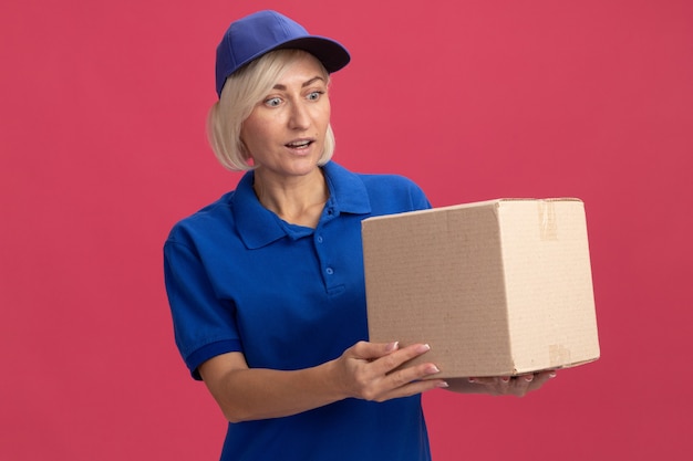 Donna bionda di mezza età impressionata in uniforme blu e berretto che tiene e guarda una scatola di cartone isolata sulla parete rosa
