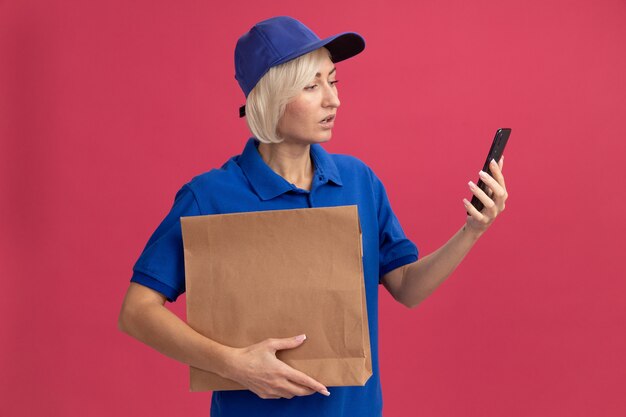 Impressed middle-aged blonde delivery woman in blue uniform and cap holding paper package and mobile phone looking at phone isolated on pink wall