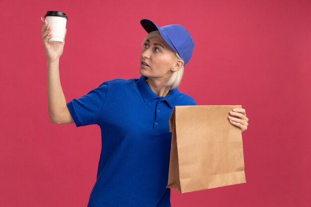 青い制服を着た印象的な中年の金髪の配達の女性とコーヒーカップを見てプラスチック製のコーヒーカップと紙のパッケージを保持しているキャップ