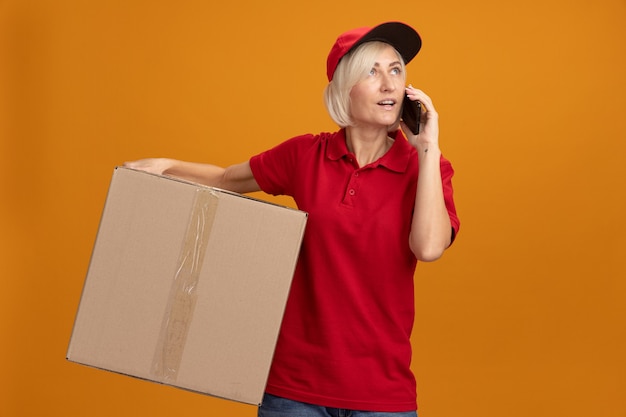 Impressed middle-aged blonde delivery woman in red uniform and cap holding cardboard box talking on phone looking up isolated on orange wall with copy space