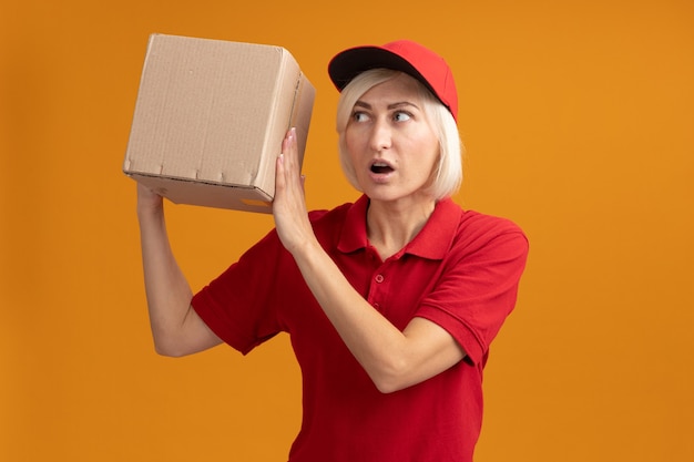 Impressed middle-aged blonde delivery woman in red uniform and cap holding and looking at cardboard box isolated on orange wall with copy space