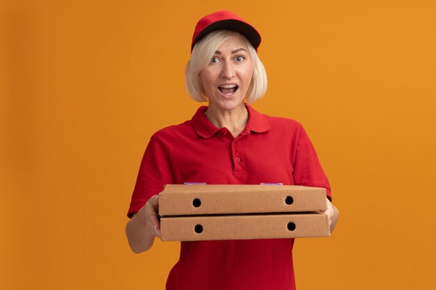 Impressed middle-aged blonde delivery woman in red uniform and cap holding pizza packages looking at front isolated on orange wall with copy space