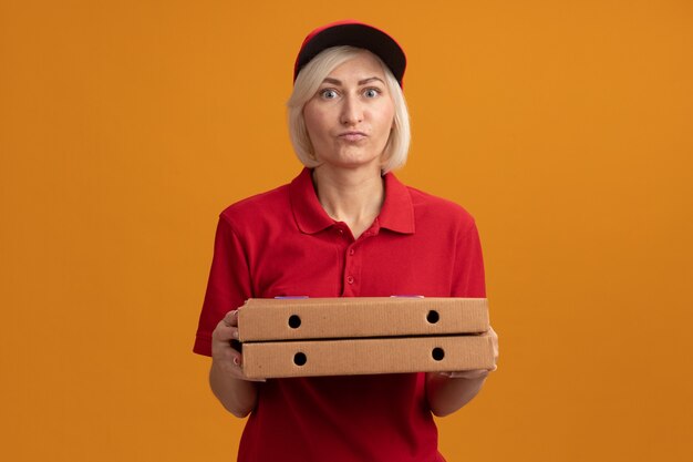 Impressed middle-aged blonde delivery woman in red uniform and cap holding pizza packages looking at front isolated on orange wall with copy space