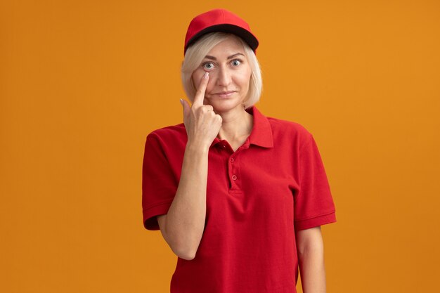 Impressed middle-aged blonde delivery woman in red uniform and cap pulling down eye lid 
