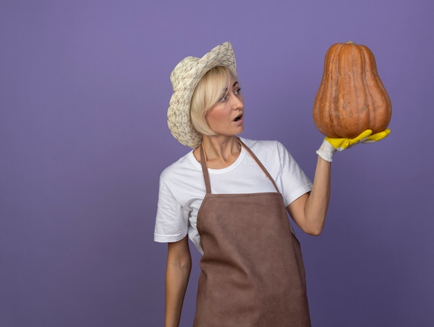 Free photo impressed middle-aged blonde gardener woman in uniform wearing hat and gardening gloves holding and looking at butternut pumpkin