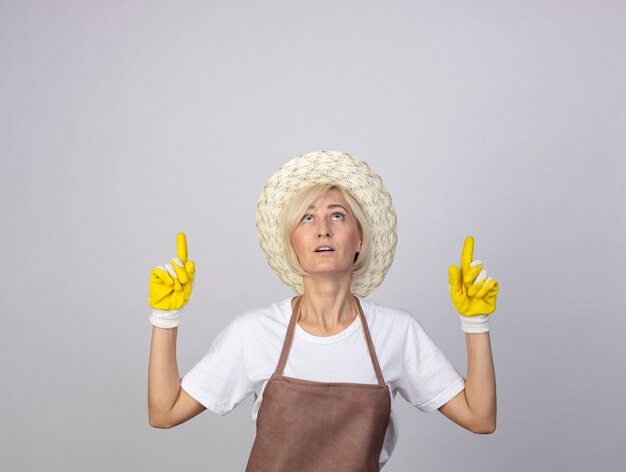 Impressed middle-aged blonde gardener woman in uniform wearing hat and gardening gloves looking and pointing up 