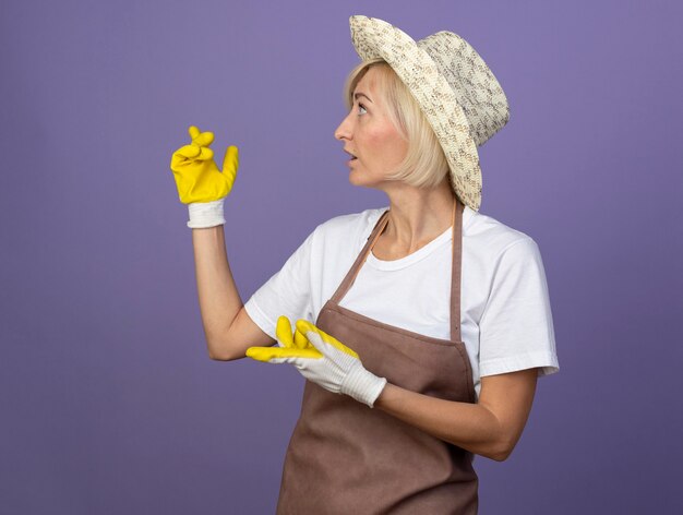 Впечатленная блондинка-садовница средних лет в униформе в шляпе и садовых перчатках, смотрящая в сторону, указывающую позади, изолированную на фиолетовой стене с копией пространства