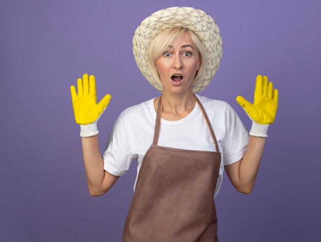 Donna bionda di mezza età impressionata del giardiniere in uniforme che indossa cappello e guanti da giardinaggio che mostrano le mani vuote