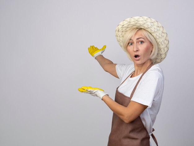 впечатленная блондинка среднего возраста женщина-садовник в униформе в шляпе и садовых перчатках, стоящая в профиль и смотрящая вперед, указывая руками за спину
