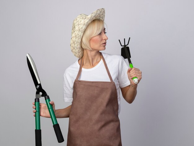 Впечатленная блондинка-садовница средних лет в униформе в шляпе с ножницами для живой изгороди и граблями смотрит в сторону