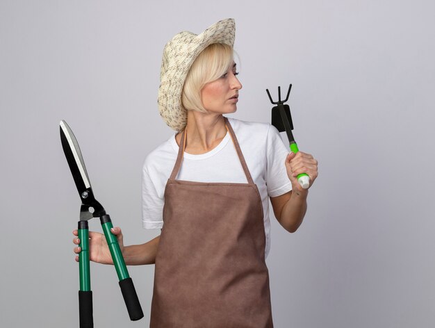 Impressed middle-aged blonde gardener woman in uniform wearing hat holding hedge shears and hoe-rake looking at side 