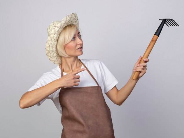 Donna bionda di mezza età impressionata del giardiniere in uniforme che indossa il cappello che tiene guardando e indicando il rastrello